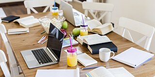 Auf dem Tisch liegen diverse Arbeitsmittel, Laptops, Bücher, Schreibblöcke, Bücher und Stifte sowie Getränke, Popcorn und Äpfel.