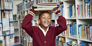 A girl is in a library and carries a pile of books above her head. 
