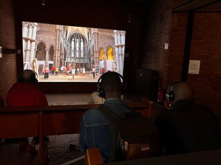 Besucher sitzen mit Kopfhörern in einem Ausstellungsraum und betrachten eine großformatige Videoprojektion einer gotischen Kirche.