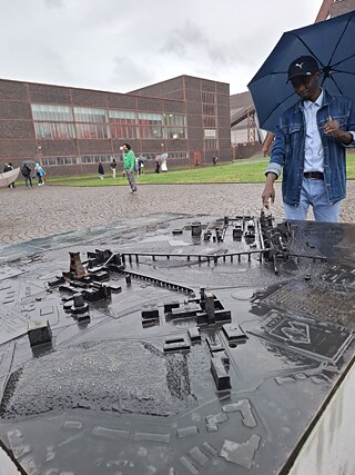 Person mit geöffnetem Regenschirm betrachtet bei Regen ein taktiles Geländemodell auf einem Platz vor einem großen Industriegebäude. Im Hintergrund bewegen sich mehrere Besucher über das Gelände.