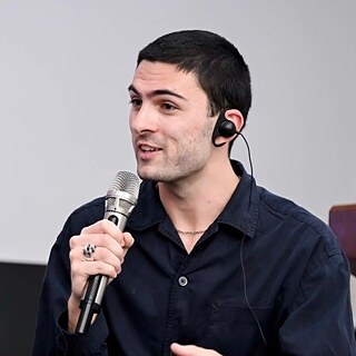 a man with dark hair and a dark shirt speaks into a microphone