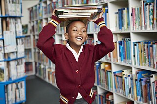 A girl is in a library and carries a pile of books above her head. 
