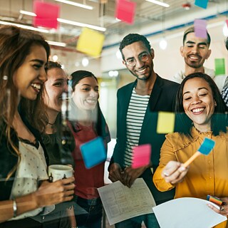 Eine Gruppe verschiedener Personen arbeiten kreativ mit Post-its an einer Glaspinnwand. Sie sind sehr konzentriert und haben Spaß.