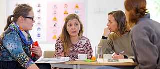Four participants are seated around a table, discussing in a workshop or seminar setting.