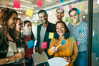 Eine Gruppe verschiedener Personen arbeiten kreativ mit Post-its an einer Glaspinnwand. Sie sind sehr konzentriert und haben Spaß.