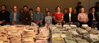 The film characters are sitting on chairs arranged in two rows. With unhappy faces they are staring in front of them. Stacked on the table in front of them are thousands of GDR-Marks.