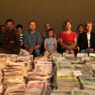 The film characters are sitting on chairs arranged in two rows. With unhappy faces they are staring in front of them. Stacked on the table in front of them are thousands of GDR-Marks.
