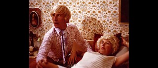 Person in patterned shirt beside a reclining person in a retro living room with floral wallpaper.