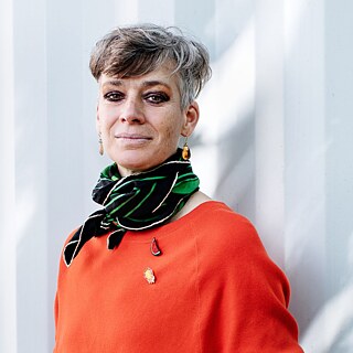 Portrait einer Person mit kurzem grauem Haar, orangefarbenem Pullover und gemustertem Halstuch vor einer hellen Architektur.