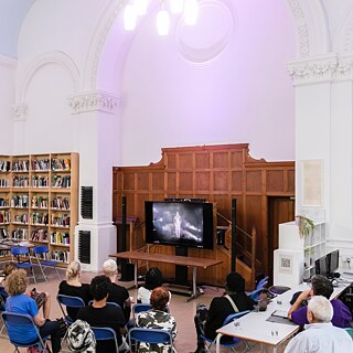Audience members seated in a library-style reading room watch a video screening on a large monitor positioned in front of a wood-panelled structure. Tall white arches, bookshelves, computers, and a large framed artwork are visible within the spacious hall.