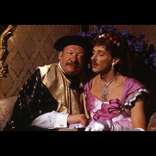 Two people in ornate theatrical costumes sit closely together in a warmly lit room with patterned wallpaper and a cabaret-like atmosphere