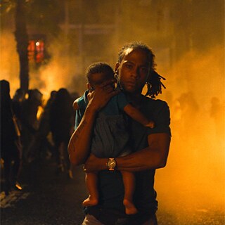 Ein Mann, mit Baby auf dem Arm und einem großen Feuer im Hintergrund.