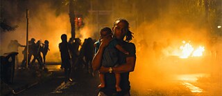 Ein Mann, mit Baby auf dem Arm und einem großen Feuer im Hintergrund.