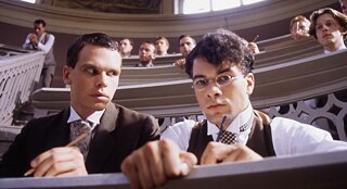 Two male students sit in a tiered lecture hall, writing at their desks while other attendees watch from the rows behind.