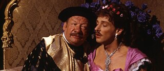 Two people in ornate theatrical costumes sit closely together in a warmly lit room with patterned wallpaper and a cabaret-like atmosphere