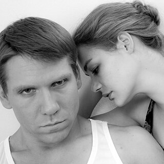 Black and white image of a grim looking man and a woman at his side who leans her forhead against the side of his head.