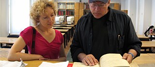 A man and a woman standing behind a table are looking at a book