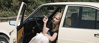 Colour image of a woman with long blond hair sitting at the front of a car with the door open and smoking a cigarette
