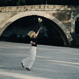 Colour image of a woman with long blond hair who jumps up in the air against the background of an old bridge with a round arch