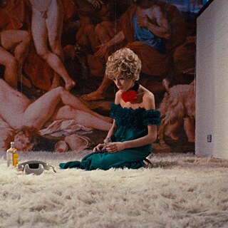 A woman with reddish hair in a green dress and a big red rose decorating her neck is sitting on a fluffy carpet looking dispondent. 