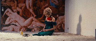 A woman with reddish hair in a green dress and a big red rose decorating her neck is sitting on a fluffy carpet looking dispondent. 