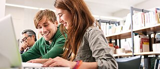 A young man and a young woman sit in the library in front of a laptop.