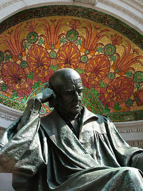 Four large bronze bas-relief panels on the walls surrounding the statue depict Hahnemann as a student surrounded by books, a chemist in the laboratory, a teacher in the lecture room, and a physician at the bedside. Hahnemann is seated on a pedestal centered in front of a curving wall. The pedestal bears the well-known principle of homeopathy, expressed in the Latin phrase 'similia similibus curantur' (like cures the like). The words 'Die milde Macht ist gross' (gentle power is great) are carved on the base of the memorial. (2000)