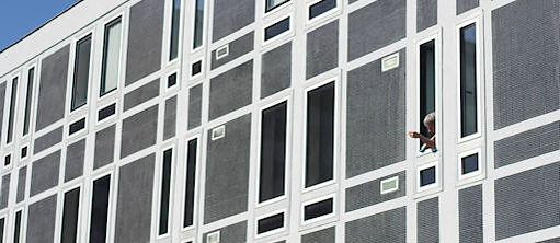 Façade d'une barre d' immeuble gris et blanc, une femme plaisse dépasser son bras par la seule fenêtre ouverte. Tout en haut à gauche de la photo un minuscule coin de ciel bleu