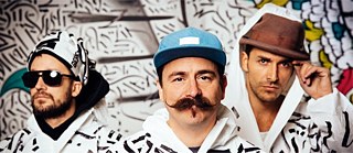 Drei Bandmitglieder in fantasievoller Kleidung vor buntem Hintergrund