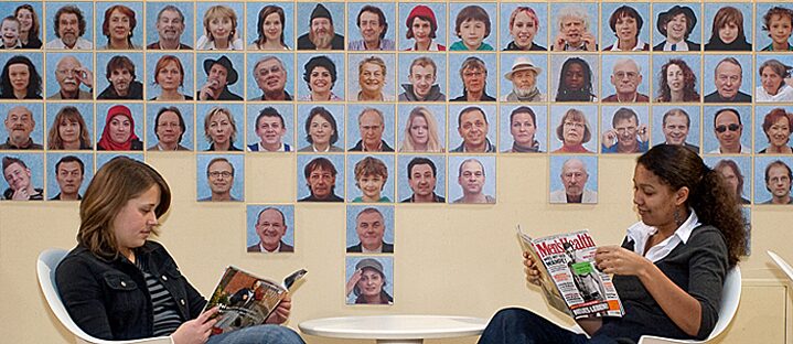 Twee vrouwen zitten aan een tafel en lezen in tijdschriften, aan de muur hangen vele portretfoto's
