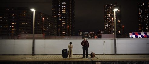Big & Little, film still: a young boy and an old man stand waiting on a train station at night, there are high rise buildings in the background.
