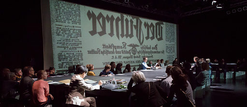 Plusieurs personnes autour d'une grande table pendant qu'un texte en allemand est projeté sur la mur.
