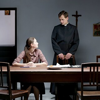 Ein Mädchen sitzt an einem Tisch und redet mit einem Priester