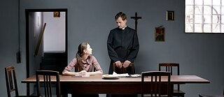Ein Mädchen sitzt an einem Tisch und redet mit einem Priester
