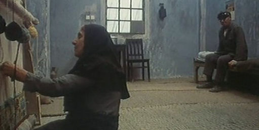 Eine iranische Frau kniet vor einer Wand, ihr Mann steht im Hintergrund