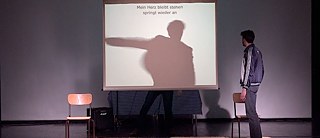 Sur une scène de théâtre, un homme regarde un écran de projection qui affiche l’ombre d’un autre homme et une phrase allemande.