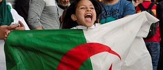 A young girl standing in a crowd, shouting and holding the Algerian flag.