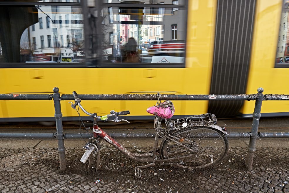 Ein Fahrradgerippe steht angeschlossen an einem Geländer, im Hintergrund fährt eine Straßenbahn.