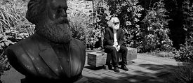 Ein Mann, der Karl Marx ähnlich sieht, sitzt neben einer Büste von Marx auf einer Bank. 