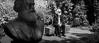 Een man die op Karl Marx lijkt, zit op een bankje naast een borstbeeld van Marx. 