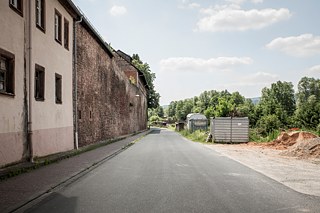Von der Schlossallee des im Main-Spessart-Kreis gelegenen Burgsinn schaut man auf die unterhalb des Schlosses gelegene alte Mauer und zerfallende Häuser. Auf der anderen Straßenseite, versteckt hinter Bäumen und Büschen, fährt die Bahn.