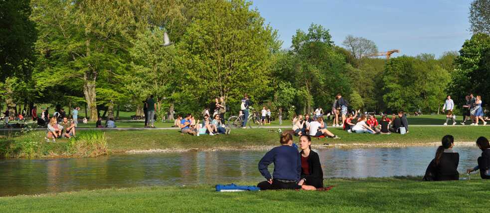 Menschengruppen sitzen an einem Bach im Englischen Garten. 