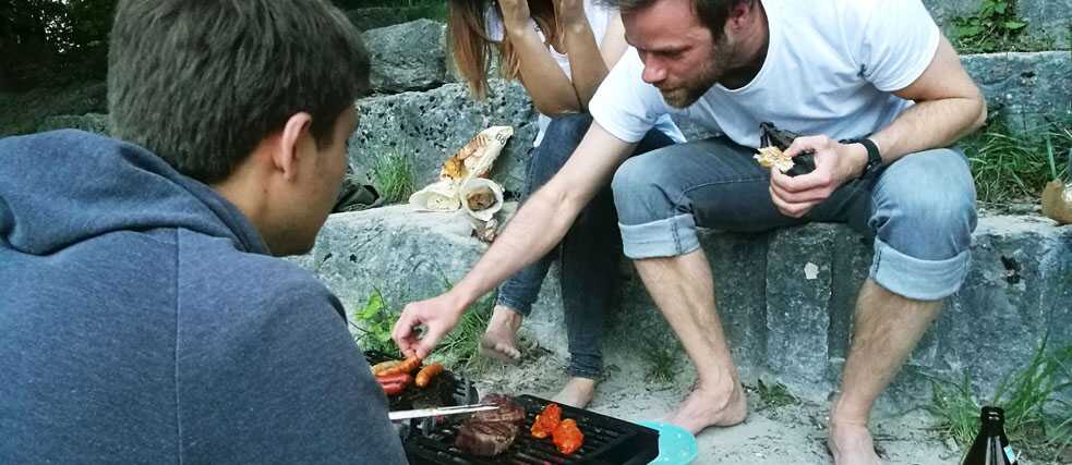 Junge Menschen sitzen um einen Grill und grillen Fleisch.