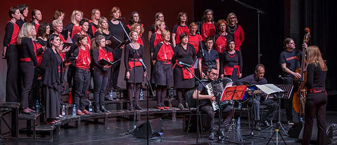 Frauenchor Kombinat steht auf der Bühne; ca. 25 Sängerinnen sind in Schwarz mit Rot gekleidet. 3 Musiker spielen vor dem Chor auf ihren Instrumenten.