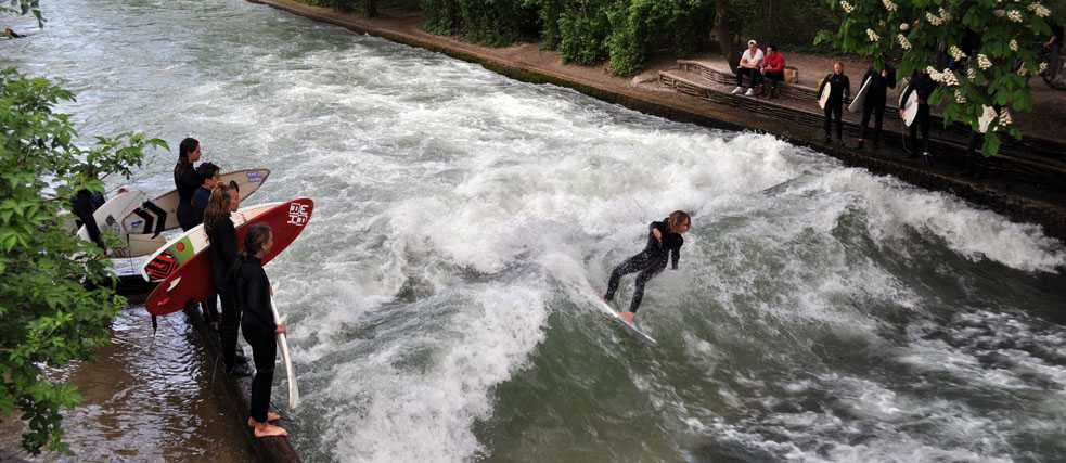 Ein Surfer surft auf einer Welle, andere Surfer warten und sehen ihm dabei zu.