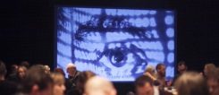 A big eye projected onto a screen observes the participants at a conference