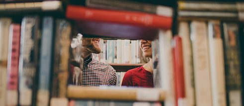 Door een spleet in een boekenkast lachen een mannengezicht en een vrouwengezicht elkaar toe.