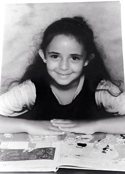 Black and white photo of the author, Huda Al-Jundi, as a child with long black hair at primary school with an open book in front of her.