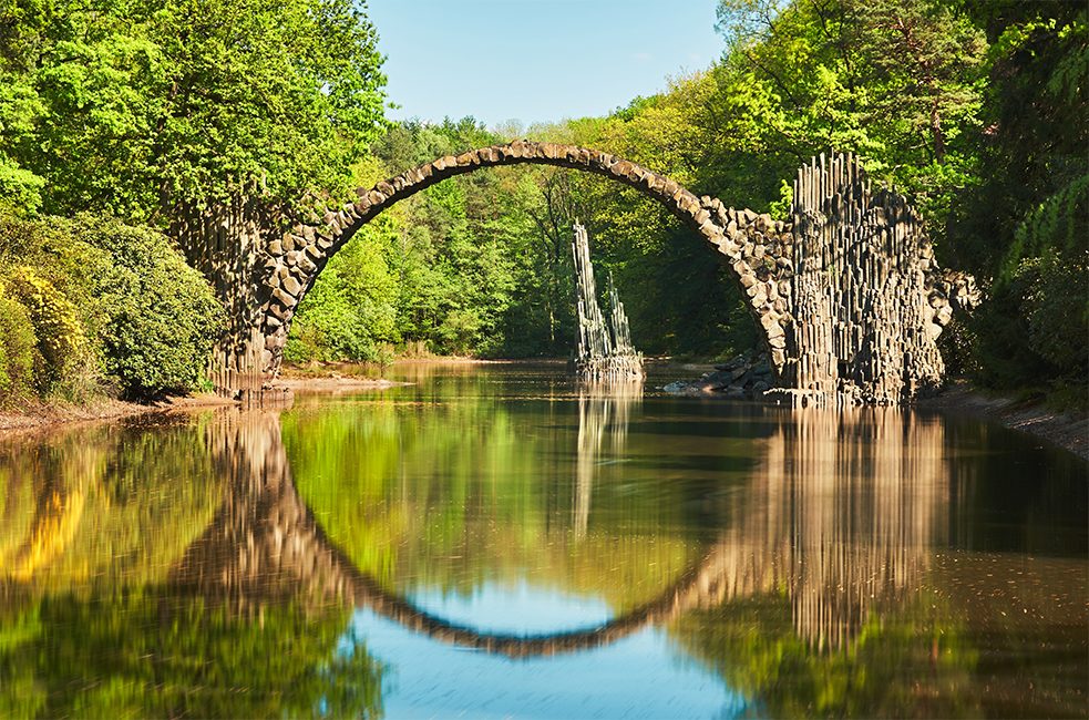 <b>Міст Ракоц: ідеальне відображення</b><br/>Немов створений для фотографій міст Ракоц у ландшафтному парку Кромлау в Саксонії. У народі його називають чортовим мостом. Збудований у 1882 році, він знаходиться у найбільшому в Німеччині заповіднику рододендронів. Його відображення у водах озера Ракоц утворює з мостом ідеальну геометрію кола у казковій атмосфері базальтових колон, що підносяться з води.