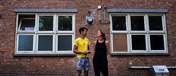 Een man en een vrouw staan voor een bakstenen gebouw en kijken elkaar glimlachend aan. 